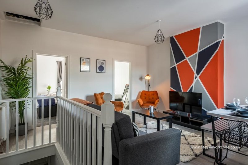 Modern open-plan living area with orange accent wall, seating, TV, and geometric artwork