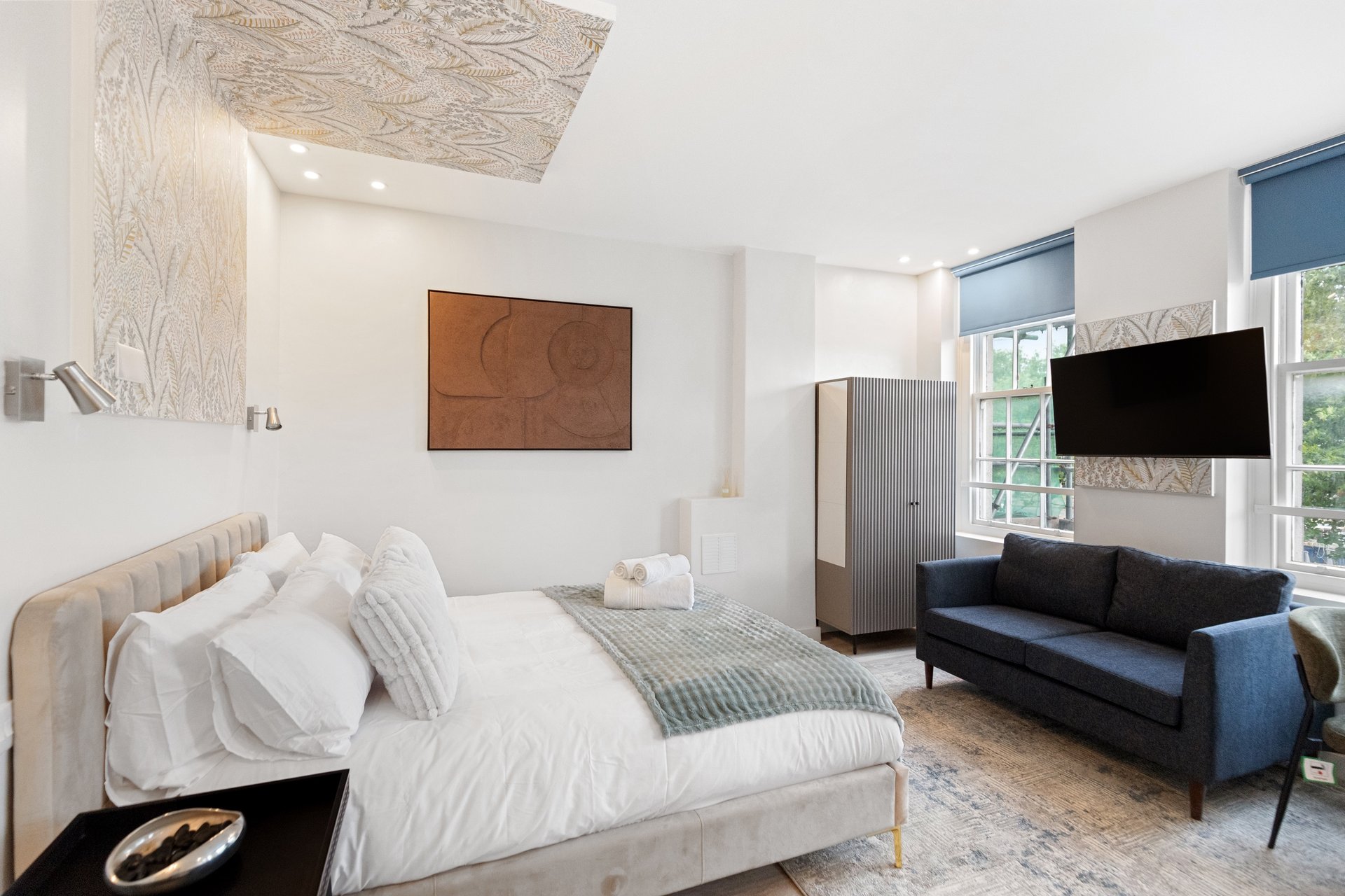 Bedroom with upholstered bed, white bedding, wooden artwork, sofa seating area and wall-mounted TV