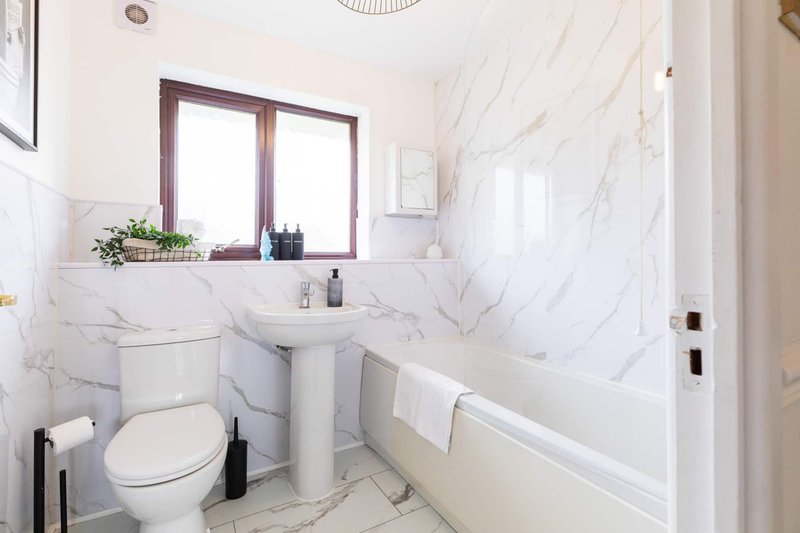 Bright bathroom with marble-effect tiles, pedestal sink, toilet, and window with burgundy frame