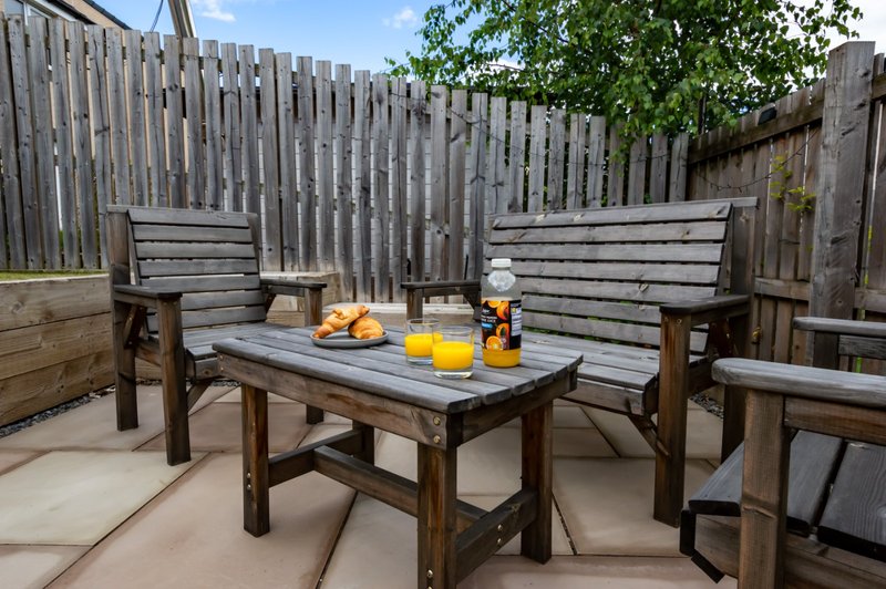 Outdoor patio with wooden benches, table, and garden furniture under blue sky