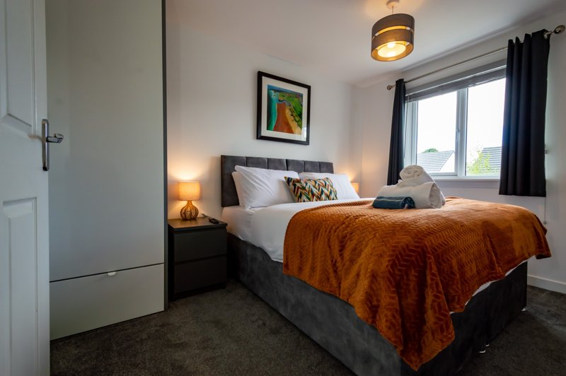 Bedroom with double bed, orange throw, bedside lamps, framed art and window with black curtains