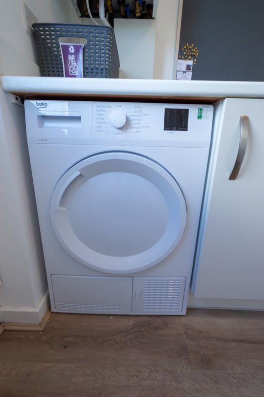 White tumble dryer next to kitchen cabinet in utility room with wooden flooring