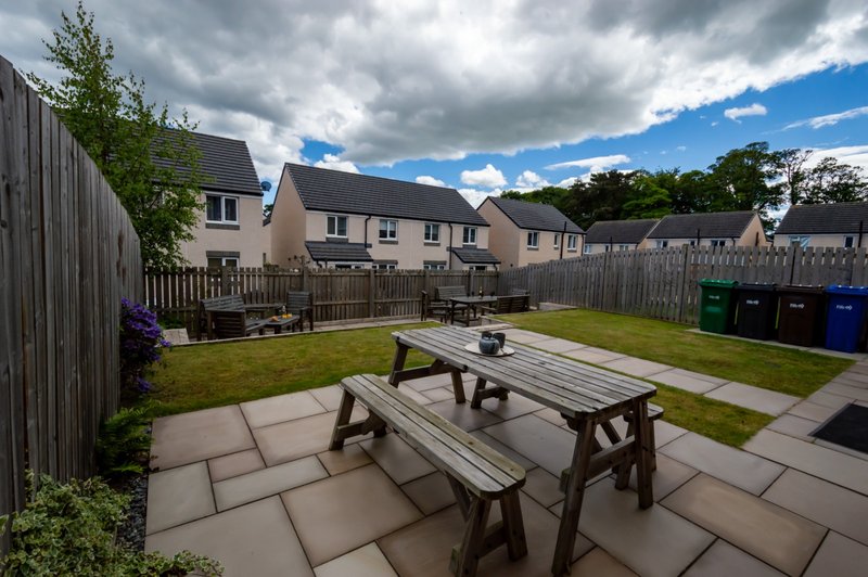 Paved garden patio with wooden picnic tables, lawn, and neighboring houses behind wooden fence