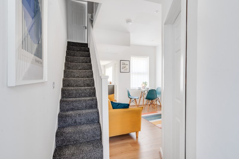 Bright hallway with gray carpeted stairs, white walls, yellow sofa and dining chairs visible ahead