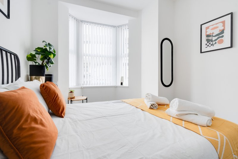 Modern bedroom with double bed, orange pillows, potted plant, and bright window with vertical blinds