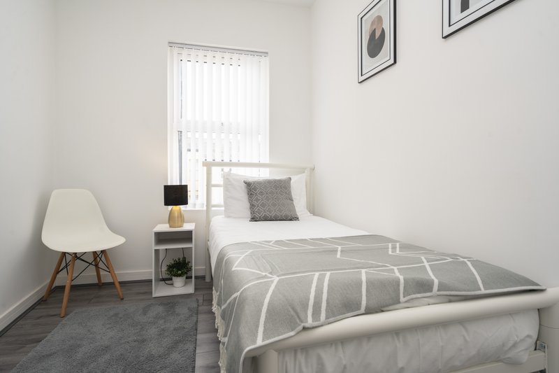 Small modern bedroom with single bed, white chair, bedside table and window with blinds