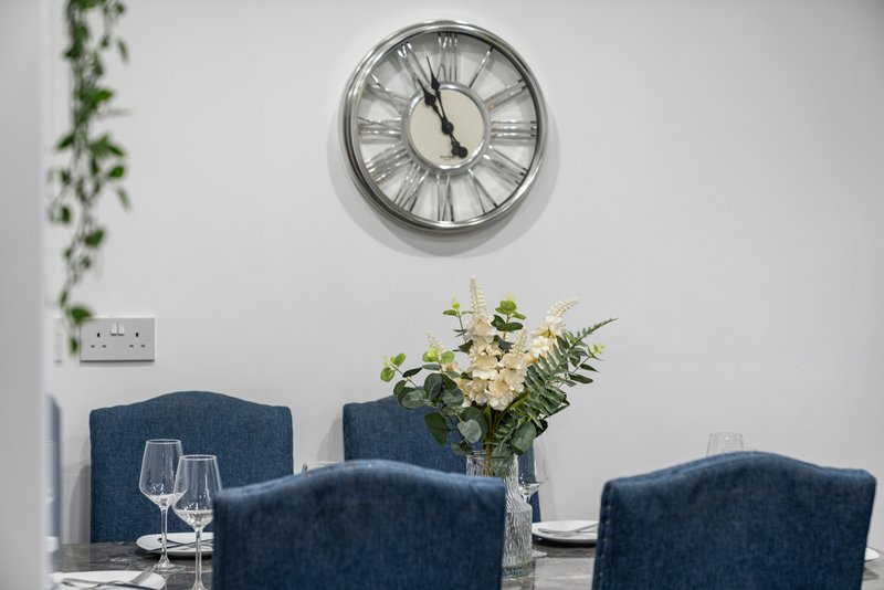 Dining table with blue upholstered chairs, wall clock, floral arrangement and glassware