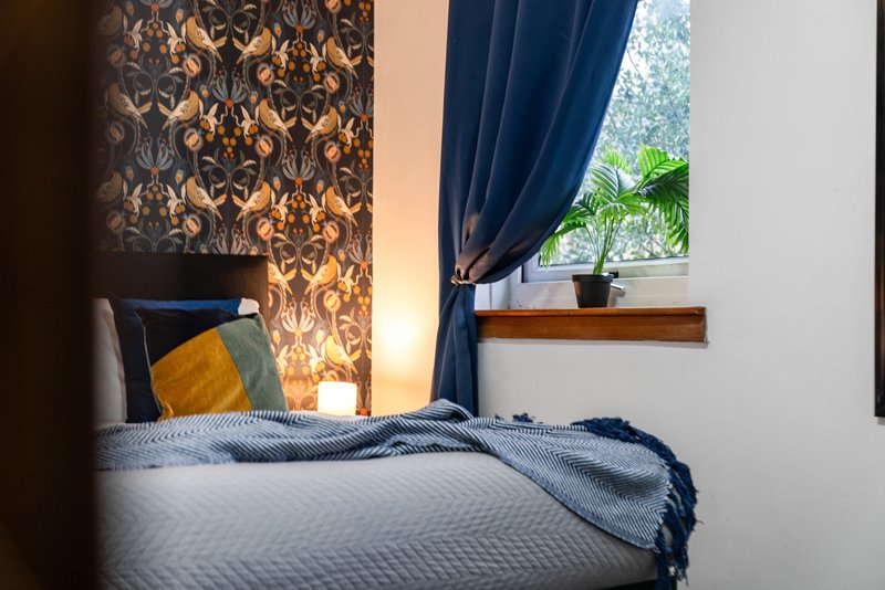 Bedroom with double bed, patterned wallpaper, blue curtains and window with plant
