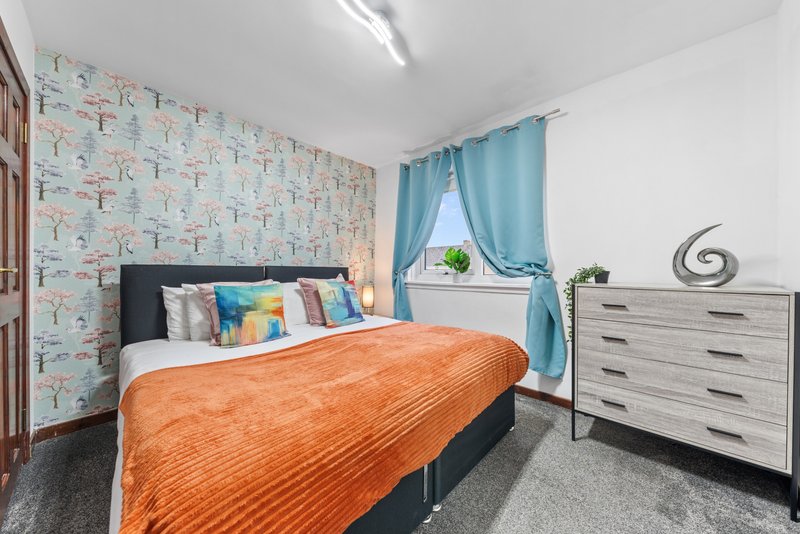 Bright bedroom with double bed, orange bedding, patterned wallpaper, blue curtains and grey dresser