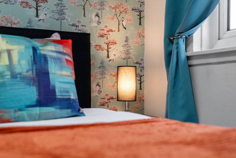 Bedroom with orange bedding, decorative tree wallpaper, blue curtains and wall lamp