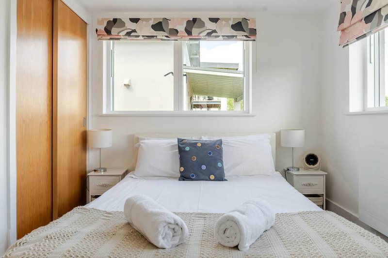Bright bedroom with double bed, white bedding, rolled towels, wooden wardrobe and patterned window valance