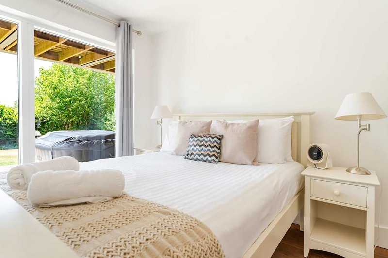 Bright modern bedroom with white bed, cream pillows, bedside lamp and patio doors to garden