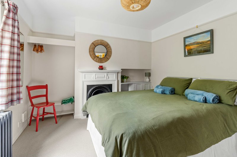 Bedroom with green bedding, red chair, fireplace, round mirror and landscape artwork