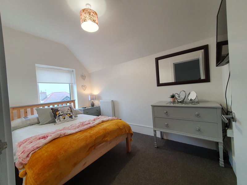Bright bedroom with wooden bed, grey dresser, mirror, and window with coastal view