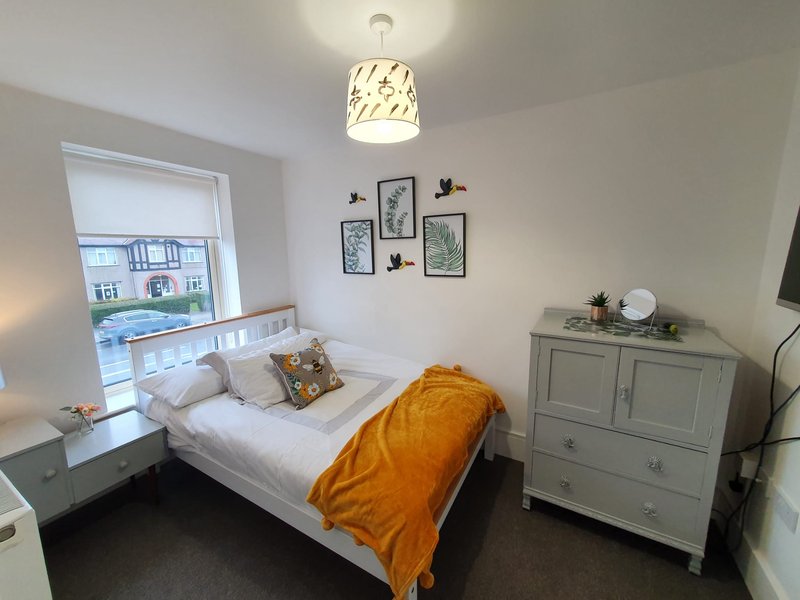 Bright bedroom with white bed, orange throw, wooden dresser, and window overlooking street