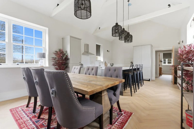 Open-plan dining and kitchen area with wooden table, grey upholstered chairs, pendant lights and large windows