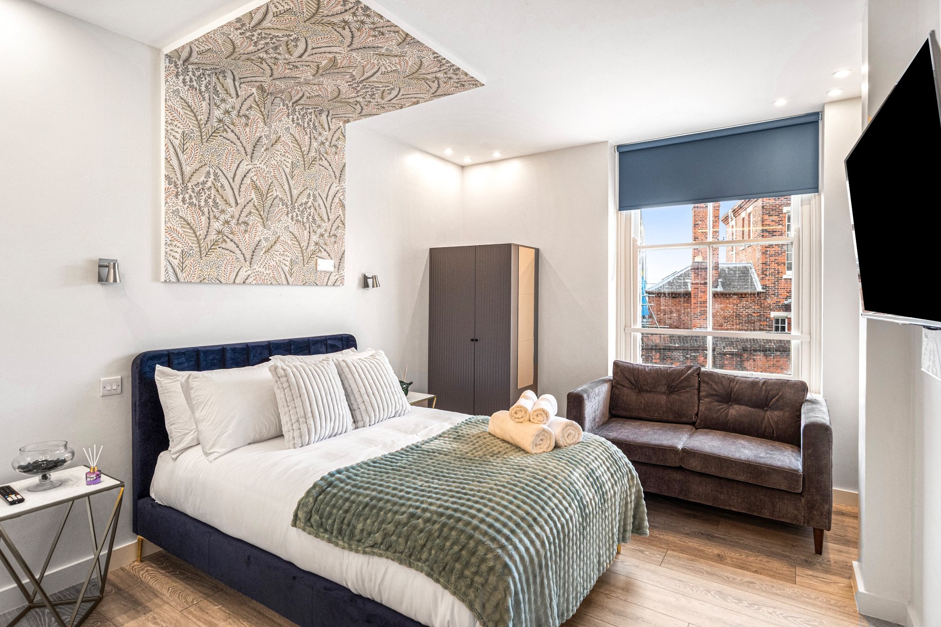 Modern bedroom with navy bed, patterned headboard, brown sofa and city views