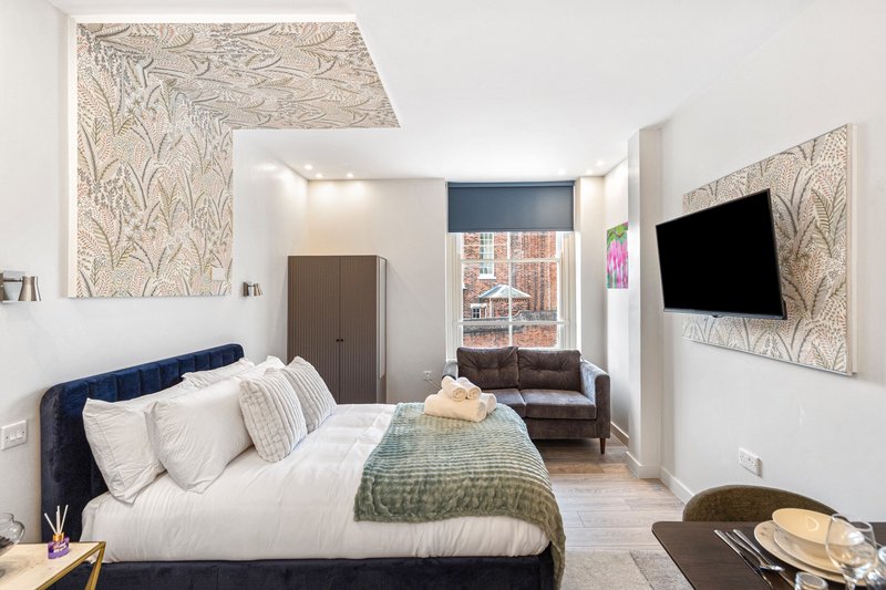 Modern studio bedroom with navy bed, white pillows, patterned wallpaper, TV, and lounge area