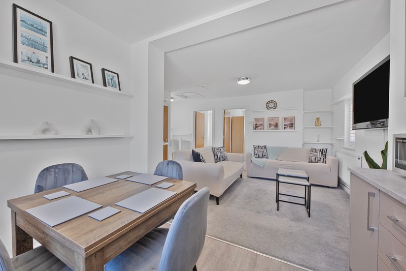 Open-plan living space with dining table, sofa, TV, and kitchen area, white walls