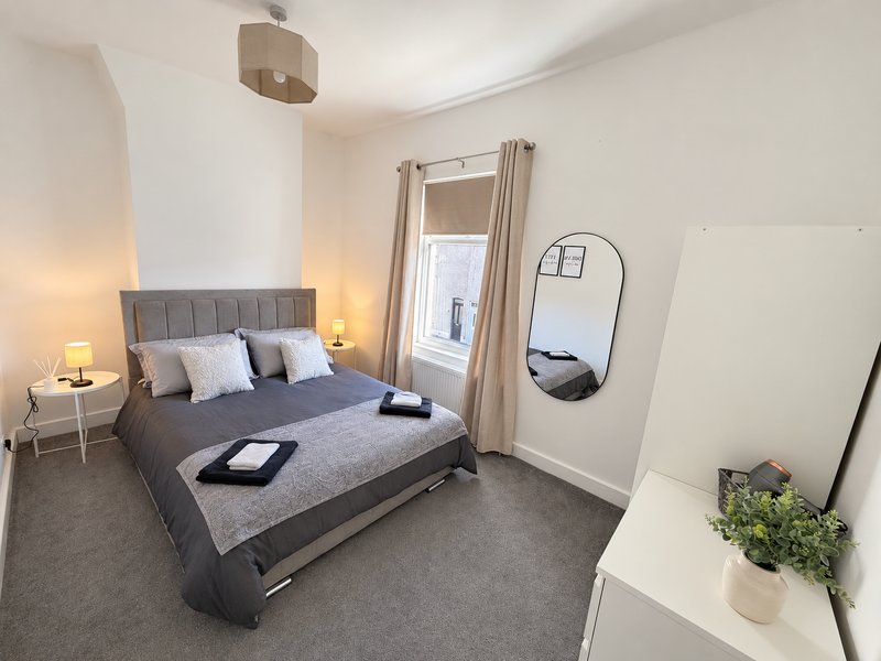 Bright bedroom with grey upholstered bed, bedside lamps, mirror, and potted plant on dresser