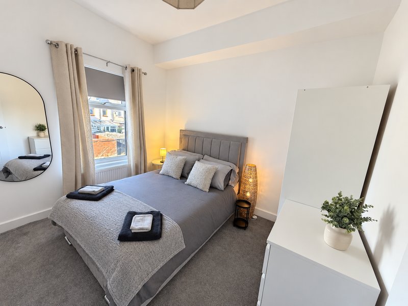 Modern bedroom with gray upholstered bed, bedside lamps, round mirror, and window with city view