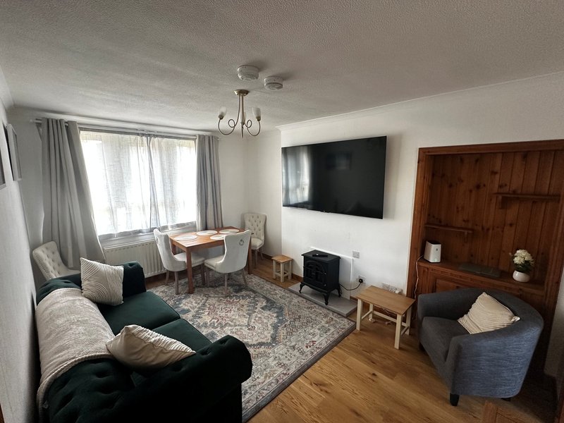 Living room with sofa and tub armchair, TV, dining table & chairs and large front facing windows.