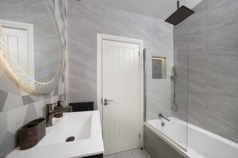 Modern bathroom with white sink, mirror, bathtub with shower enclosure and grey tile walls