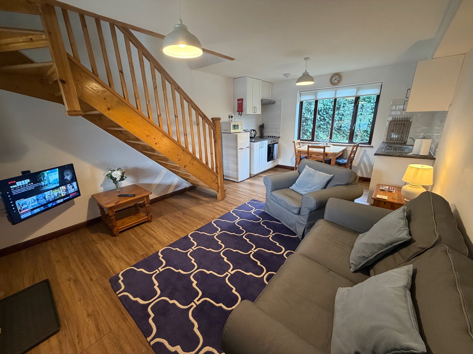Open-plan living room with wooden staircase, grey sofas, TV, and kitchen area