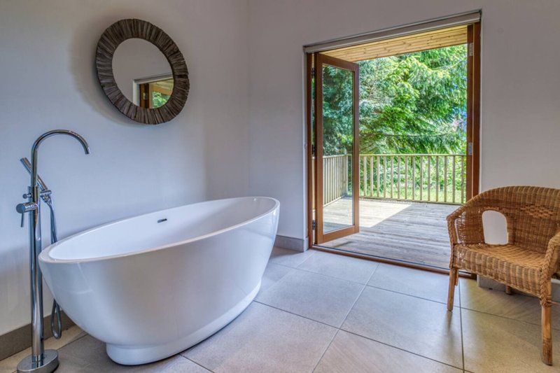 Luxury bathroom with freestanding white bath, wicker chair, round mirror and glass doors opening to balcony