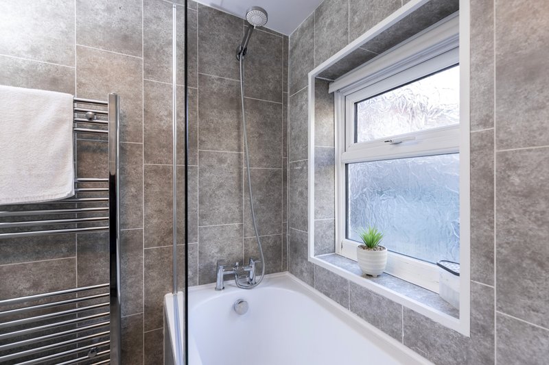 Modern bathroom with gray tile walls, white bathtub, rainfall showerhead and window with garden view