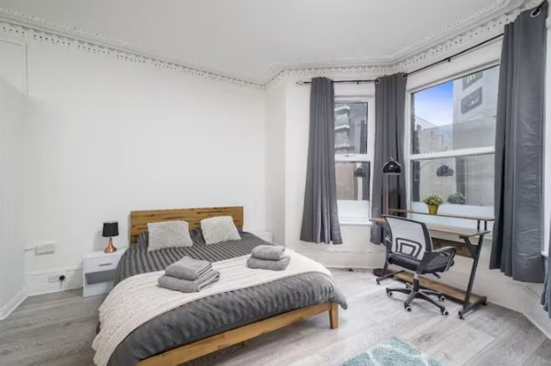 Bright bedroom with wooden bed, grey bedding, desk, office chair and large windows