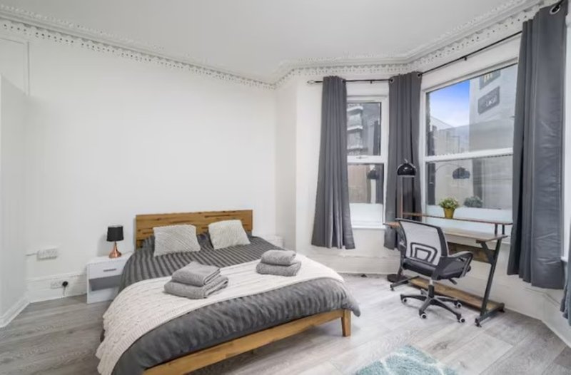 Modern bedroom with wooden bed, grey bedding, desk chair and large windows with city views