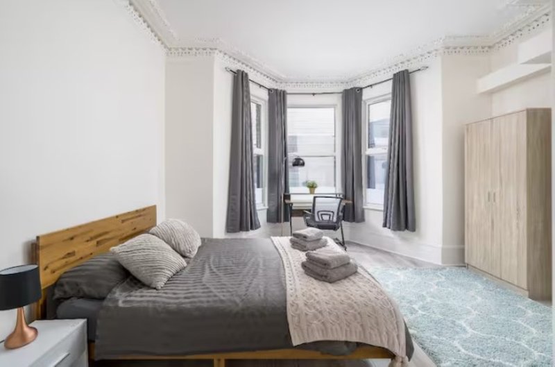 Bright bedroom with wooden bed frame, grey bedding, bay windows with grey curtains, and wooden wardrobe