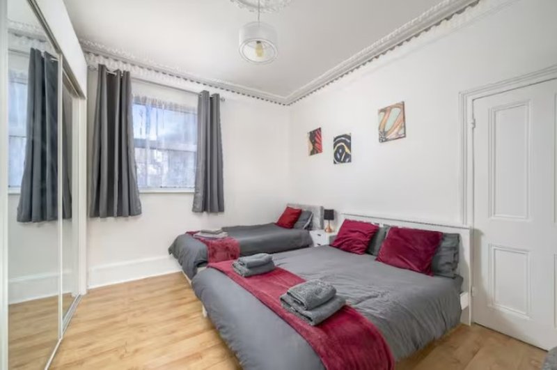 Bright bedroom with two beds, red and grey bedding, wooden floor and curtained window