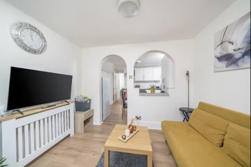 Open-plan living room with yellow sofa, TV on cabinet, wooden floor and archway to kitchen