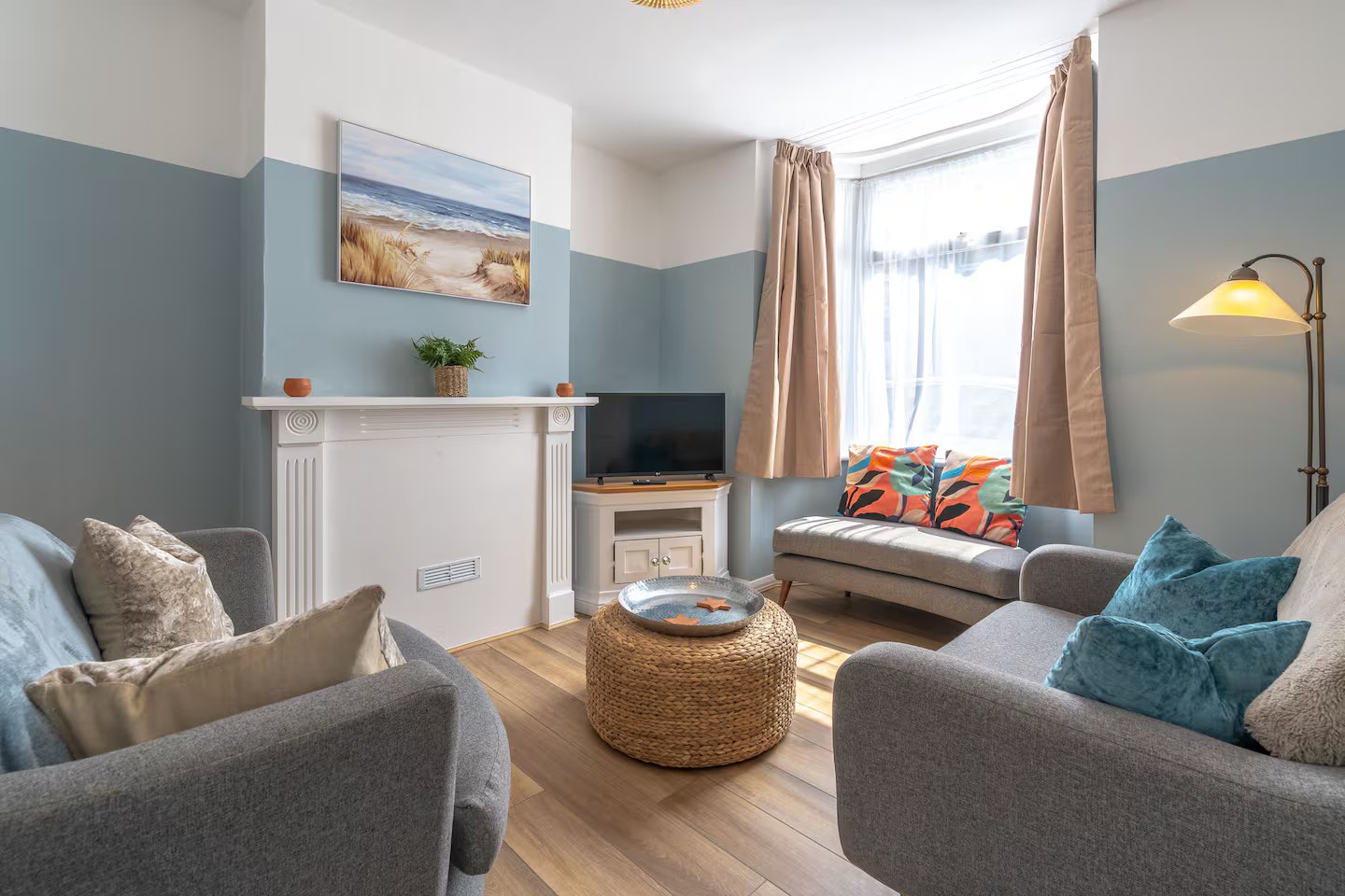 Bright living room with two grey armchairs, sofa, woven ottoman table, and coastal wall art