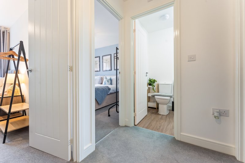 Hallway with open doors showing bedroom and bathroom, wooden flooring and white walls