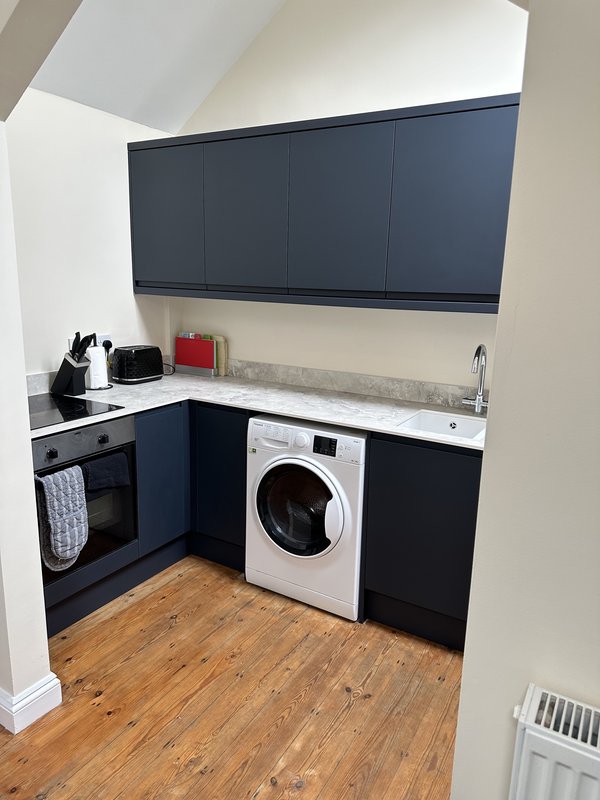 Modern compact kitchen with dark cabinetry, white washing machine, electric oven and wooden flooring