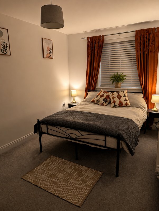 Modern bedroom with metal bed frame, white bedding, decorative cushions, pendant lamp, and orange curtains