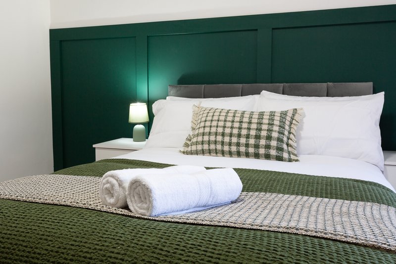 Modern bedroom with dark green panelled headboard, white pillows, rolled towels and green bedding