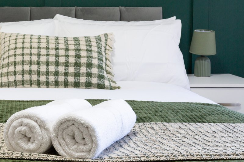 Bedroom with made bed, green gingham pillow, white pillows, rolled towels, and bedside lamp
