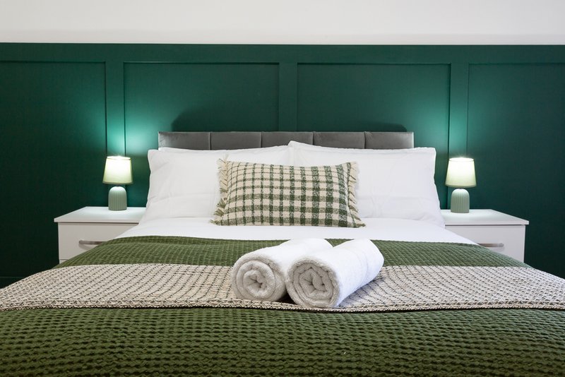 Modern bedroom with green headboard, white pillows, rolled towels on bed, and bedside lamps