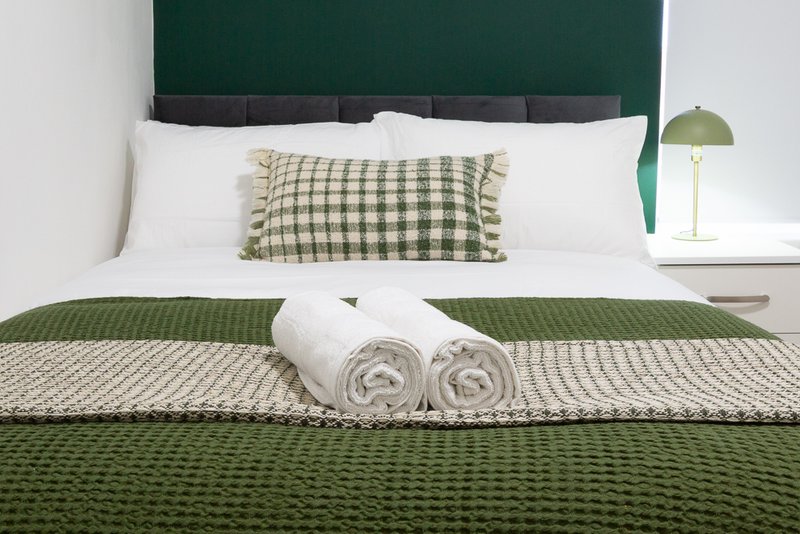 Bedroom with green upholstered headboard, white pillows, checkered cushion, rolled towels, and layered bedding
