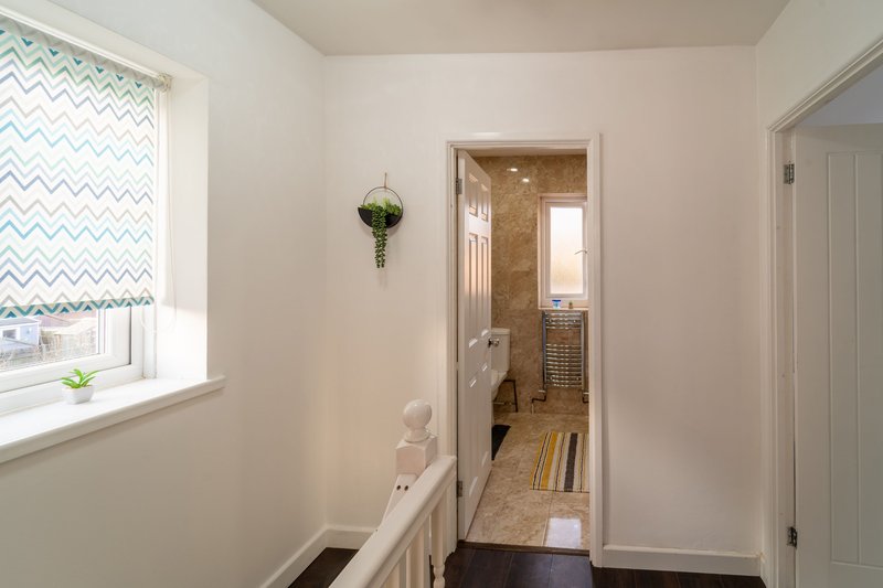 Bright hallway with white walls, window with patterned blind, hanging plant, and open doorway to bathroom