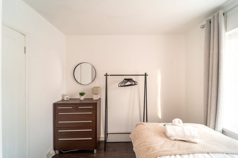 Bright bedroom with double bed, dark wooden dresser, round mirror, clothing rack and curtained window