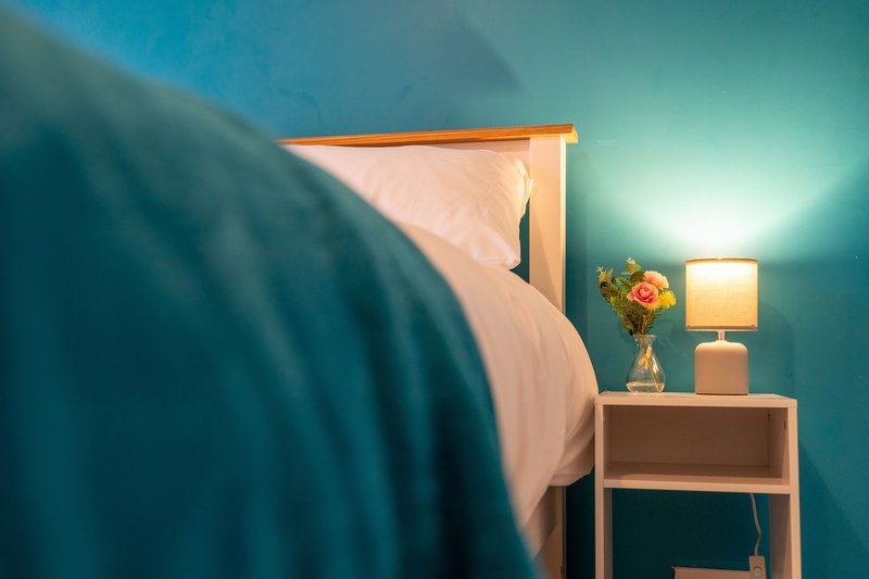 Bedroom corner with teal walls, wooden bed frame, white bedding, and wooden nightstand with lamp and flowers