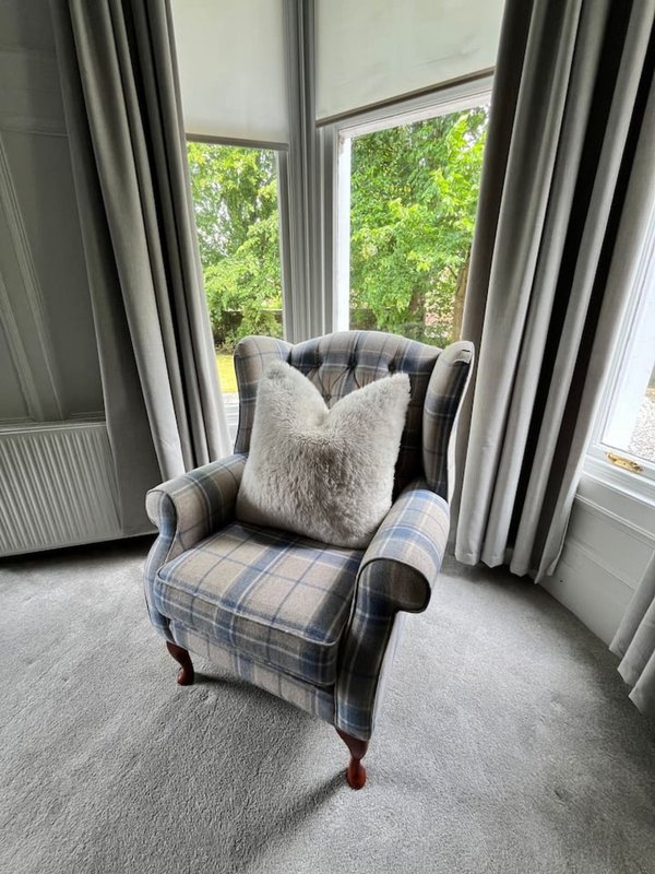 Upholstered wingback armchair with plaid fabric and cream cushion near windows with grey curtains