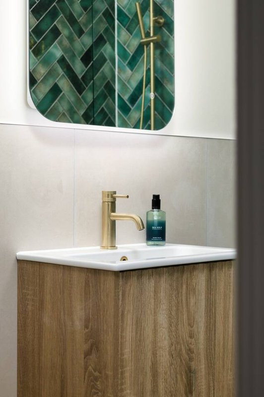 Modern bathroom sink with brass faucet, green herringbone tile mirror, and toiletries