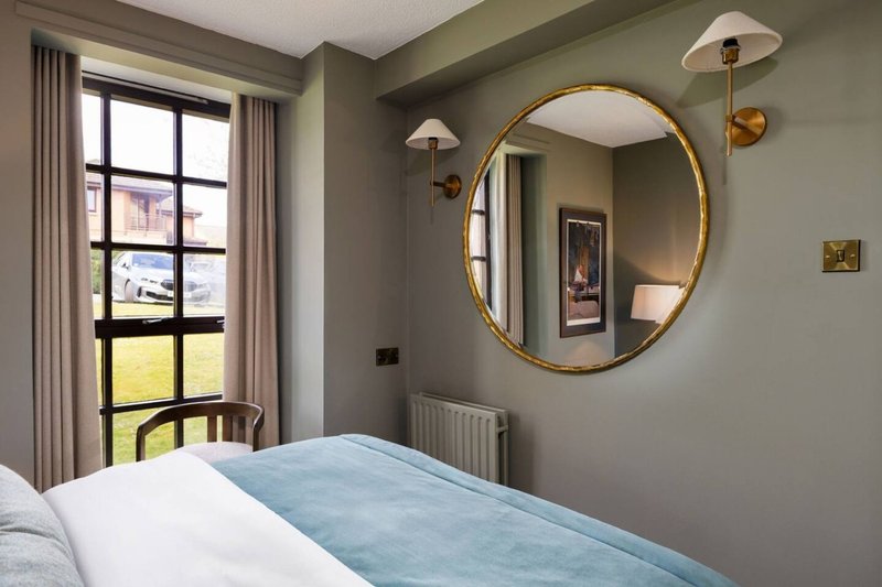 Modern bedroom with double bed, round gold mirror, and view through large window to garden