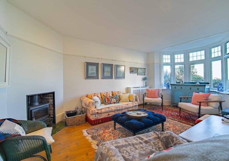 Bright living room with fireplace, patterned sofa, blue ottoman and large windows overlooking garden